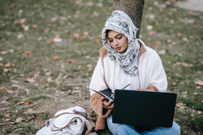 jeune femme voilée adossée à un arbre, qui écrit sur sa tablette